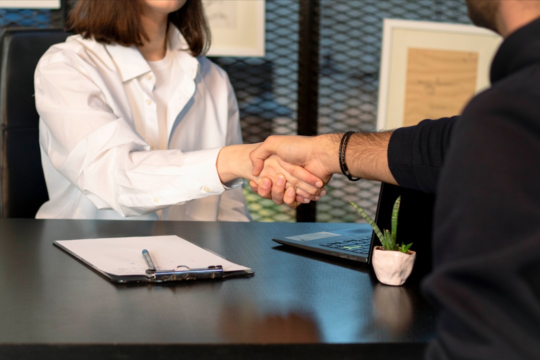 Two people shaking hands across a table in front of a laptop after reviewing documents