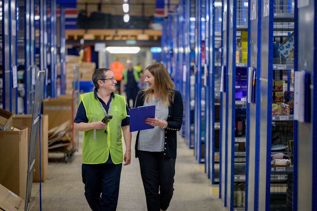 Two colleagues walking and talking in a warehouse aisle, one holding a clipboard to review supplier stock